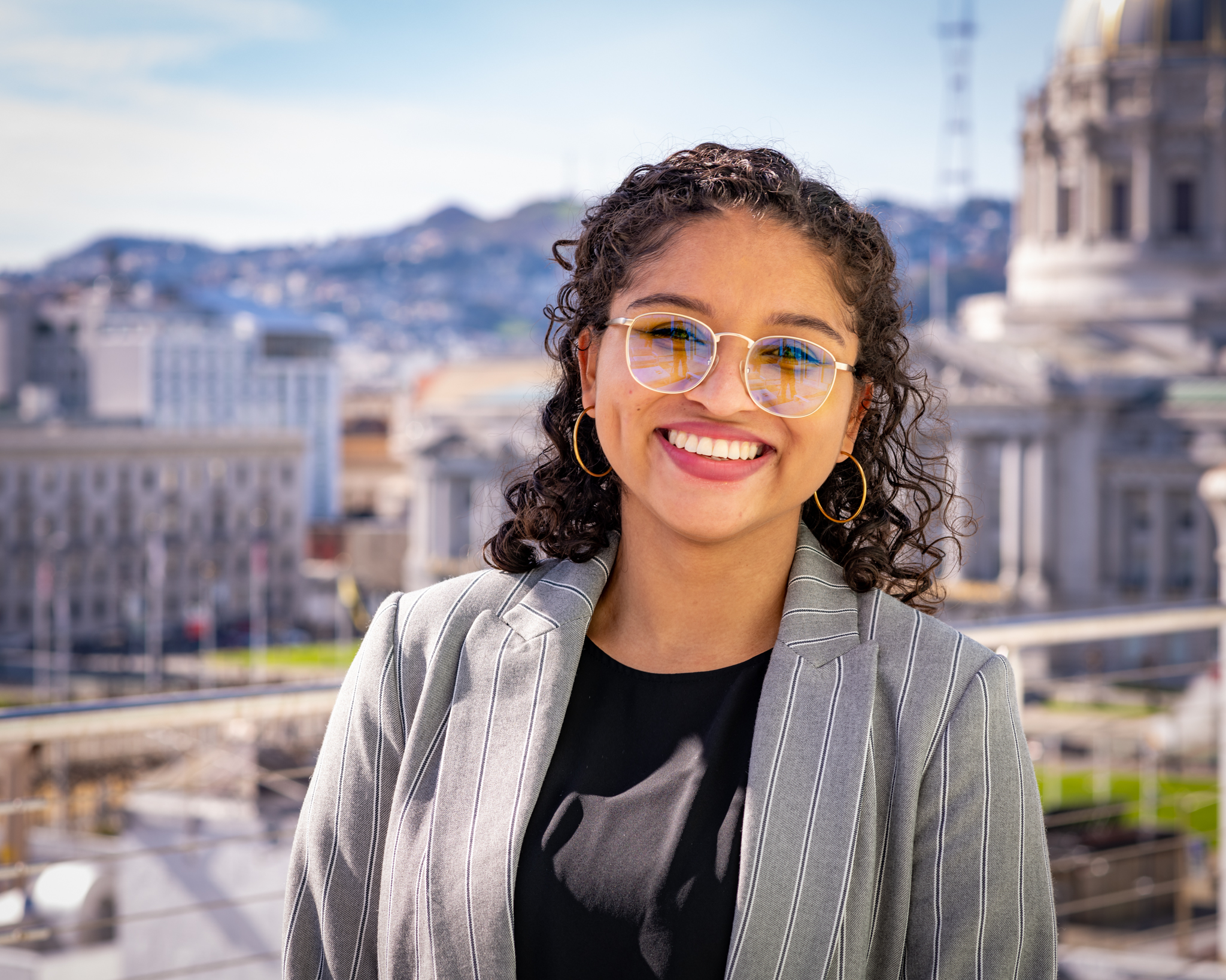 anne-rodriguez-webb Hispanic woman with long, curly brown hair, wearing a gray suit jacket and black shirt, smiling in front of a state or federal capital building.