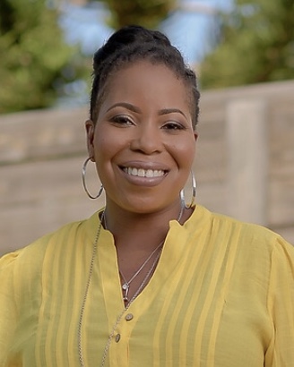 shelyna-brown Black woman with tied-back, black hair, wearing a yellow button down, in front of a brown fence and green tree.