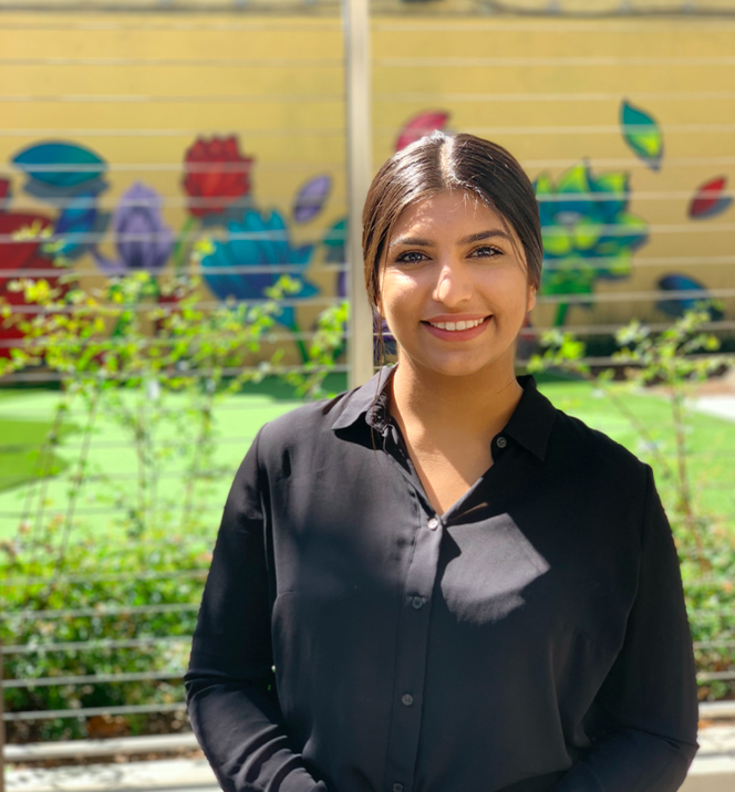 Sukhjit Middle Eastern woman with black hair in a ponytail, wearing a black button down shirt, smiling in front of a painted elementary school wall.
