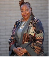 Virginia Jones Black woman with dark brown hair, wearing an orange and brown, patterned suit jacket, smiling in front of a gray brick wall.